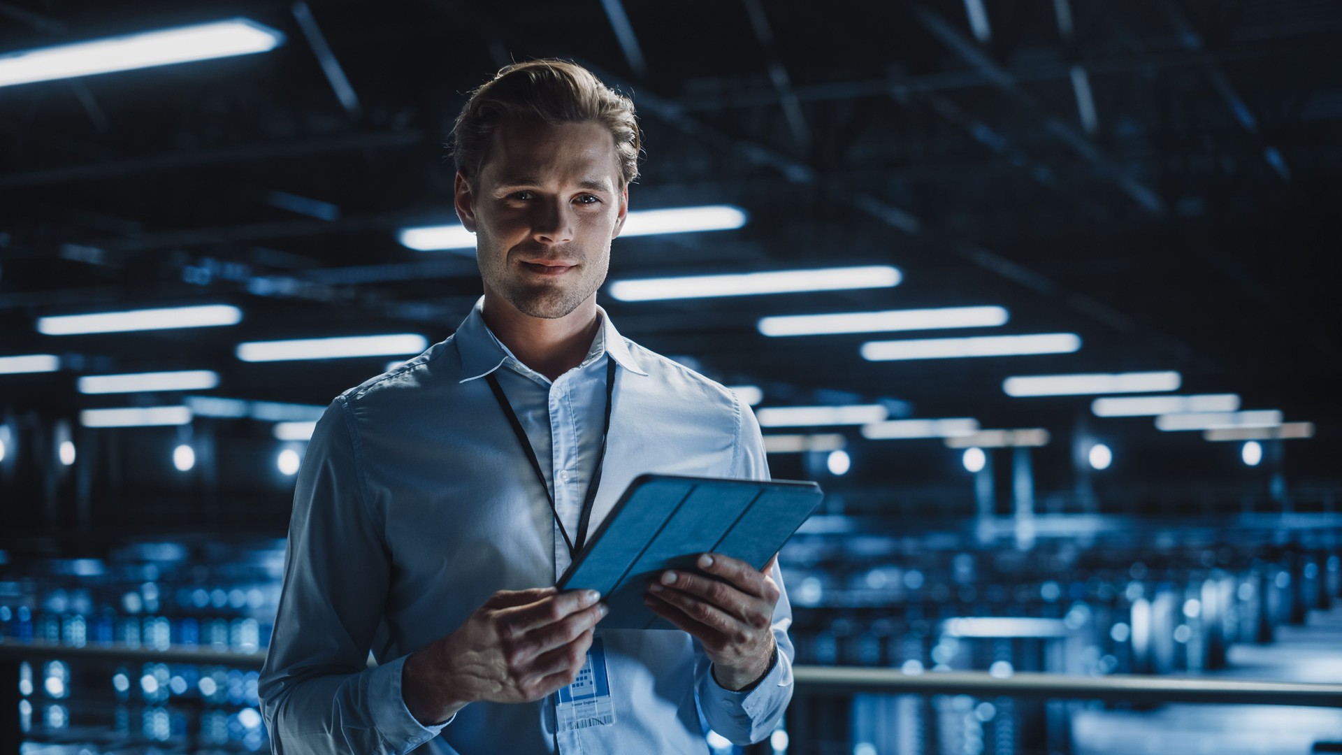 Portrait of Handsome Smiling IT Specialist Using Tablet Computer in Data Center, Looking at Camera. Succesful Male e-Business Engineer Working in Big Server Farm Cloud Computing Facility.